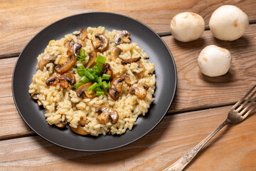 Homemade lemon risotto with champignon mushrooms on a black plate. Whole raw champignons behind. Rustic wooden table. Healthy vegan eating concept.