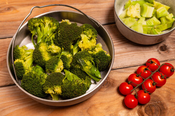 Steamed broccoli florets in a stainless-steel steamer, cooked broccoli stalk pieces in a bowl, and a sprig of cherry tomatoes.