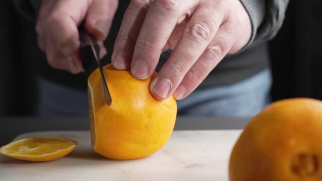 Hands slice an orange into even rounds on a cutting board with a kitchen knife. Close-up food prep shows peel texture, juicy pulp, and fresh citrus color.