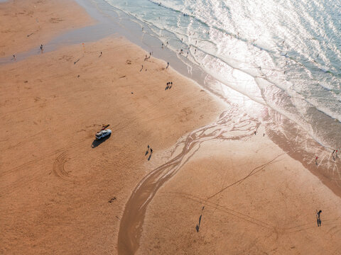Aerial view of the golden sands meeting the shimmering sea, where people stroll and a lone vehicle rests, reflecting the sunlight, Perranporth, England, United Kingdom.