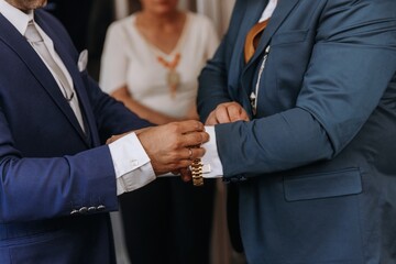 Close-up of a groom in a blue suit being helped by a best man to adjust his gold watch and cufflinks during wedding morning preparations