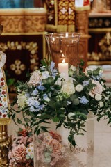 Lit white candle in a tall glass vase surrounded by a floral wreath of roses and greenery inside an Orthodox church