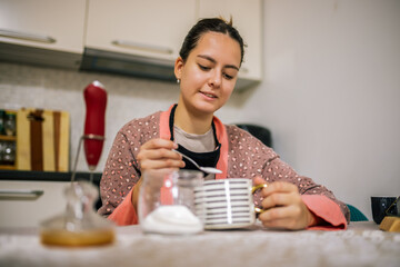 oung woman stirs her coffee with a hand whisk and adds sugar, preparing her morning drink at home.