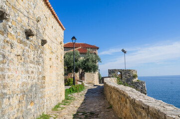  Narrow stone walkway in Ulcinj&rsquo;s Old Town, Montenegro, leading past ancient walls to a seaside lookout. The Adriatic Sea glistens under a clear blue sky beside historic fortress structures.