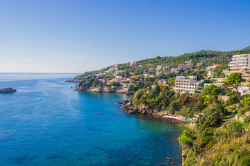  Coastal view of Ulcinj, Montenegro, with hillside houses and resorts overlooking the Adriatic Sea. Crystal-clear blue waters meet lush greenery under a bright summer sky.