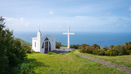 Minimalist white church on green grass, with a large cross, on a cliff in the Azores islands with...