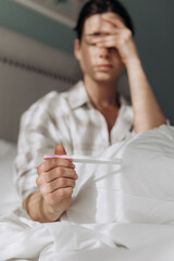 Young woman seating on bed, holding pregnancy test, displaying a concerned expression, surrounded by soft bedding, evoking feelings of anxiety and hope