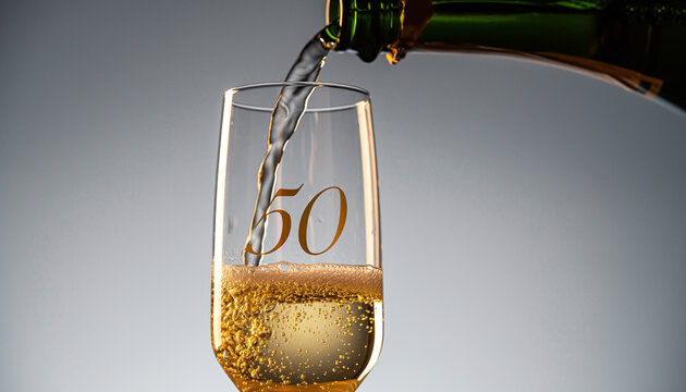 Glass engraved with the number 50. Champagne bottle fills the glass with alcohol. Celebrating milestone, anniversary or birthday - Powered by Adobe