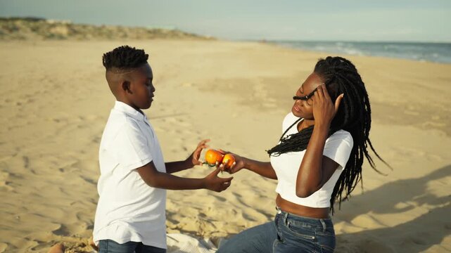 Happy african american mother with dreadlocks playfully tossing ripe oranges to delighted son as they enjoy fun beach day together, radiating warmth and family joy