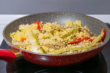 A red frying pan on a stovetop holds a colorful mix of millet, zucchini and peppers