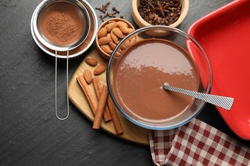 Liquid chocolate dough in bowl, spices, nuts and cocoa powder on black table, flat lay. Space for text