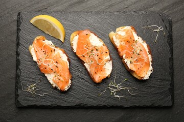 Tasty bruschettas with salmon, cream cheese, microgreens and lemon slice on black table, top view