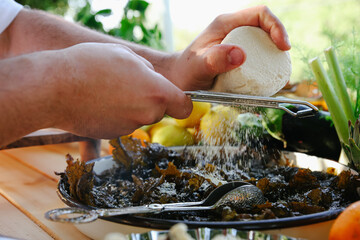 Close-up of Chef's Hands Grating Fresh White Cheese with a Zester or Grater Over a Dark Leafy Green or Cooked Vegetable Dish
