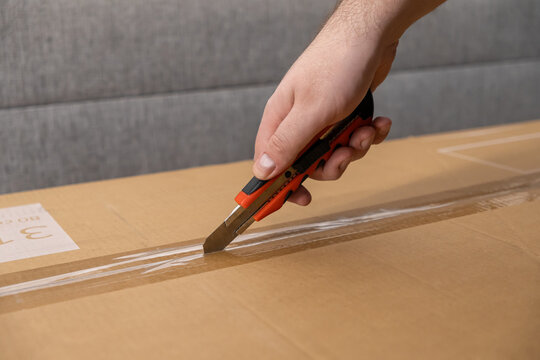 Hand using a red box cutter to slice packing tape on a brown cardboard box, symbolizing efficient parcel delivery and quick unpacking process