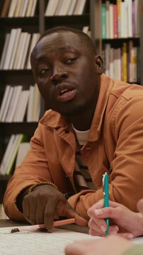Vertical shot of young African American male language tutor pointing at English irregular verbs on poster clarifying grammar rule for adult students during library based lesson