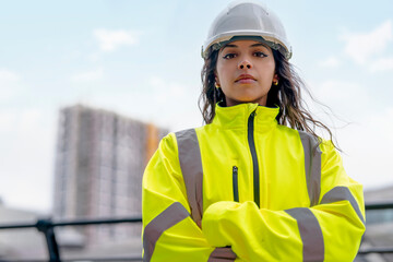 Portrait of female construction worker, wearing a reflective yellow safety jacket and helmet, stands with arms crossed, showcasing confidence at a busy construction site