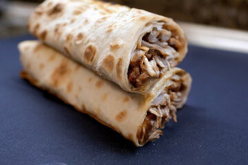 Vertical close-up of a freshly grilled shawarma or durum wrap, sliced in half diagonally and stacked, exposing the filling of meat, vegetables, and sauce, set on a dark, matte surface