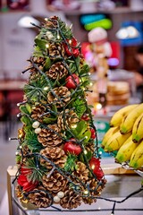 Christmas tree decoration made of pine cones, apples, and greenery, adorned with lights, set against a blurred cafe background, creating a festive atmosphere with warm ambiance