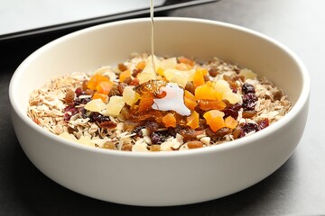 Making granola. Pouring honey into bowl with oat flakes and dried fruits on grey table, closeup