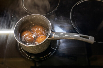 Overhead view of several brown eggs boiling in a small dark speckled saucepan on a black, modern glass induction or ceramic stovetop