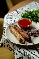 Two delicious kebabs rest on a plate, surrounded by vibrant herbs and sliced onions. A bowl of spicy sauce adds flavor, all set against a unique patterned tablecloth