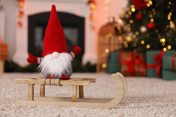 Fototapeta premium Cute Christmas gnome with sleigh on floor against blurred lights indoors, closeup