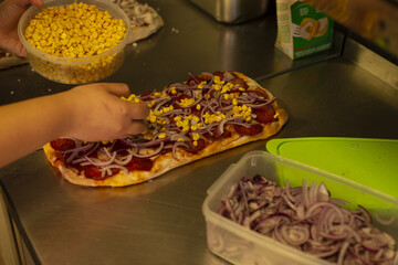 hands layering corn and onion pizza, rectangular dough topped with pepperoni and sauce, plastic tubs of ingredients on stainless