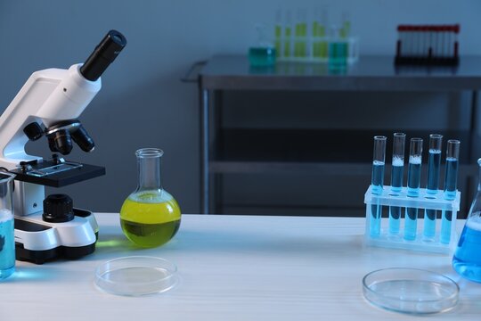Microscope and laboratory glassware with different liquids on white wooden table indoors - Powered by Adobe