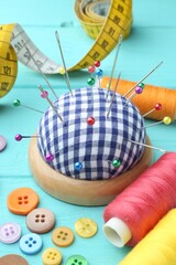 Checkered pincushion with pins, needles, buttons, measuring tape and spools of thread on light blue wooden table, closeup