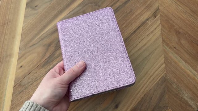 Female hand holding and displaying a purple glitter tablet or e-reader case over a wooden table. Tech accessory concept shot in 4K with natural light.
