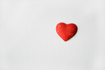 A small red heart with a few drops of water on the surface, creating a feeling of freshness and tenderness, lies on a white background. A declaration of love and feelings.