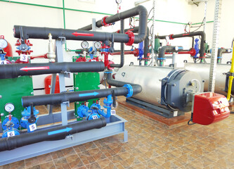 A modern boiler room with two large industrial boilers. The boilers are connected to pipes supplying water and gas. The pipes are equipped with pressure gauges, shut-off valves, pumps, and fittings.