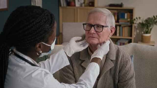 Medium close up shot of black female doctor in white coat and gloves examining lymph nodes of sick aged male Caucasian patient at home