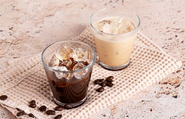 Iced coffee and black coffee served in clear glasses on beige stone table, close up