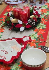 Festive Christmas table setting featuring a red floral runner, a centerpiece with red pillar candles and pinecones, and ceramic holiday serving dishes; seasonal celebration.