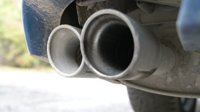 Close-up of dual car exhaust pipes at idle, emitting deep engine sound. Subtle vibrations and dust on the metal surface highlight the vehicle running at low RPM