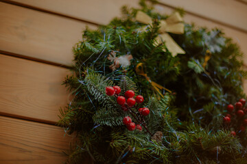 Close-up of a Christmas wreath decorated with evergreen branches, red berries, and a golden bow, hanging on a wooden wall and creating a warm festive holiday atmosphere.