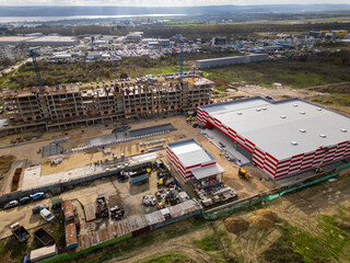 Aerial view of a large industrial building under construction with red-and-white panels, workers, equipment, and surrounding development site