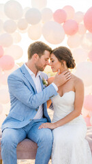 Bride in white lace gown and groom in blue suit sit close with foreheads touching. Surrounded by pastel balloons, they share an intimate moment of love and commitment on their wedding day.