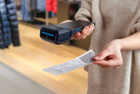 Fototapeta Hand offering printed receipt with QR code next to POS terminal inside clothing store, showing modern payment and checkout workflow