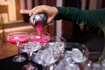 Mood setting with lively drink preparation at upscale bar, Elegant scene of bartender pouring colorful cocktail amidst festive warm lighting and guests in background