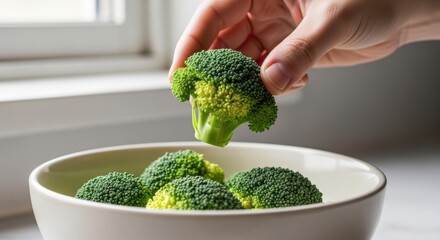 Hand holding fresh broccoli over bowl in sunlit kitchen