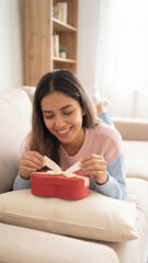 Joyful moment as a person opens a red heart-shaped gift box on a cozy couch. The warm living room setting evokes love, surprise, and celebration in a relaxed home atmosphere.