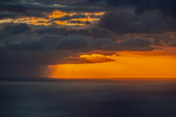 Dramatic sunset over the ocean, with dark storm clouds above and intense orange sunlight breaking through in powerful rays across the horizon