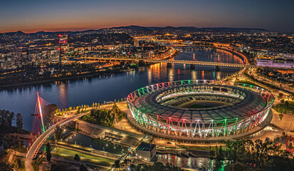 Budapest. Drone night view of a stadium and riverfront cityscape illuminated with colorful lights, showing bridges, buildings and vibrant urban life from above.