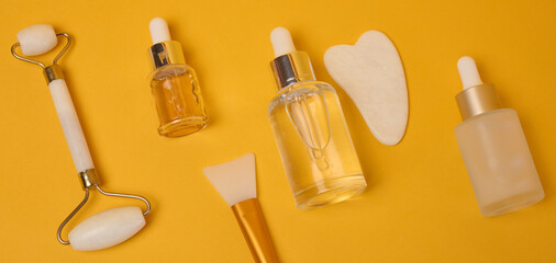 Glass bottles with serums and pipettes stand next to a massage roller and a jade gua sha scraper, a brush for applying a mask
