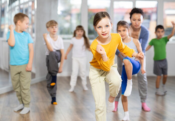 In sports studio, creative childrens team learns modern dance accompanied by experienced woman teacher. Teacher corrects movements of hands and body, instructs students to perform dance movements
