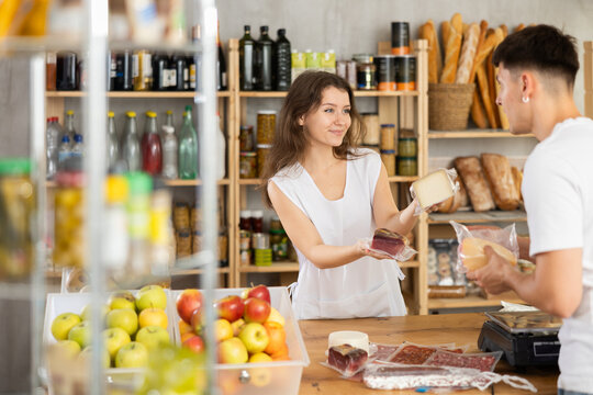 Young female seller offers cheese, dried sausage and meat to young male buyer in grocery store - Powered by Adobe