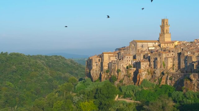 Italian town of pitigliano revealing on a tuff cliff. Picturesque medieval town of pitigliano emerging from behind a green hill, with its historic buildings built on a tuff rock cliff