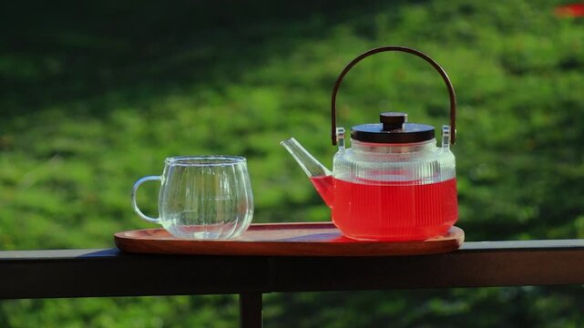 A female hand places an empty glass cup next to a hot transparent teapot filled with red hibiscus tea outdoors. Natural green background, rising steam and a calm, healthy lifestyle atmosphere.